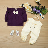 Purple top with a bow and white pants on a wooden surface with flowers and baby shoes.